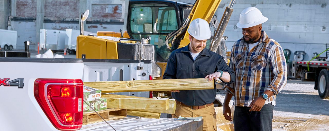 Two men in a construction zone with hard hats are pulling long wood planks off a pick-up truck.