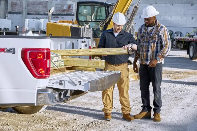 Two men wearing hardhats at a construction site removing lumber from the back of a pickup truck.